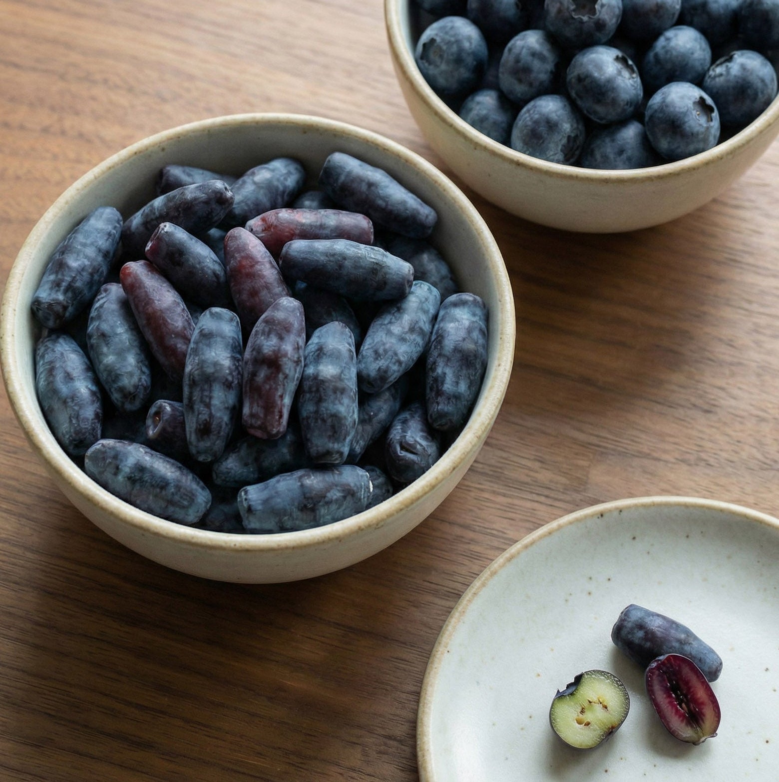 Two bowls of blueberries and haskap berries on a wooden table with a small plate of fruit.