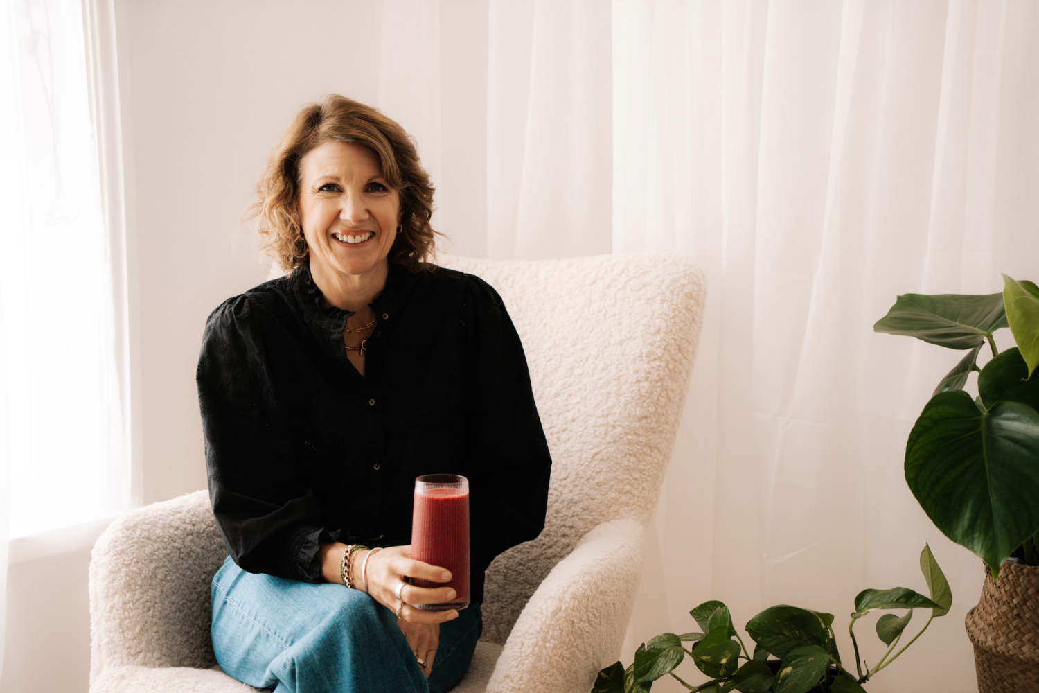 Woman sitting on a couch holding a glass of red juice in a bright room with white curtains and a plant.